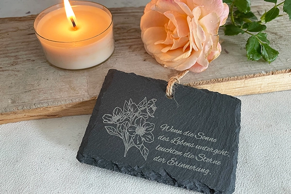 Kleine Schieferplatte mit einem gravierten Spruch und der Gravur eines Blumenstrauß. Dahinter ist ein brennendes Teelicht und eine Rose zu sehen.