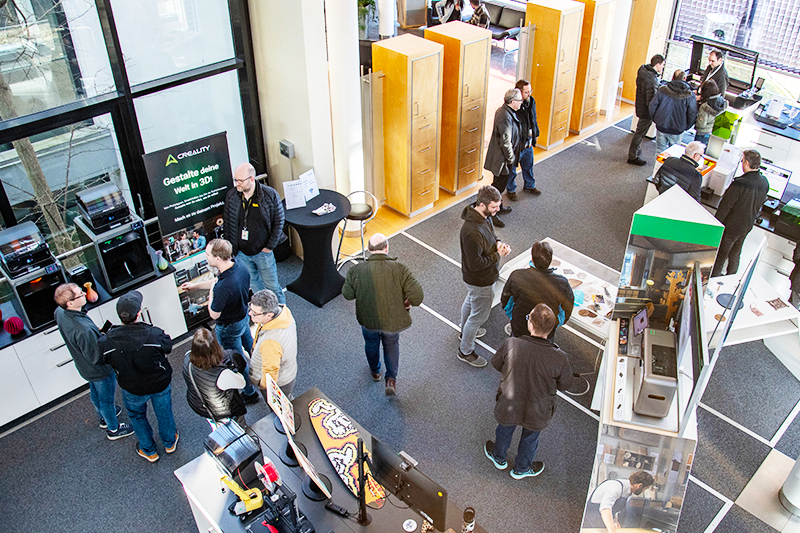 Das Bild zeigt mehrere verschiedene Menschen, die in einem Raum mit verschiedenen Lasern und 3D-Druckern stehen