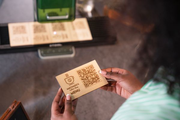 Eine Frau hält eine edle Holzkarte mit gelasertem Logo und Menü-QR-Code eines Cafe´s in den Händen.